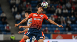 RB大宮アルディージャの市原吏音選手が、コーナーキックの場面で相手選手の上からヘディングシュートを放つ、高さと強さが分かる写真。