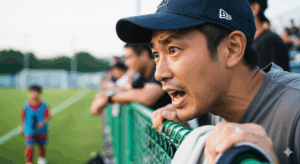 サッカーの試合中、観客席のフェンス越しに、ピッチ上の我が子へ必死に指示を送る父親の写真。