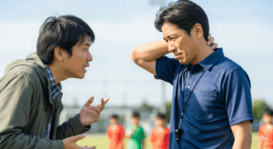 少年サッカーの試合中、保護者がコーチに対して何かを訴えかけるように話している、少し気まずそうな雰囲気の写真。